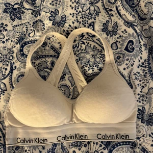 Vit sport-bh från Calvin Klein - Vit sport-bh från Calvin Klein med brottarrygg och elastiskt band med logga nedtill. Mjuk och bekväm modell i bomullsmix, perfekt för träning eller vardag. Snygg och enkel design med vadderade kupor.