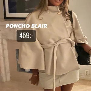 Beige poncho med bälte och knappdetalj poncho Blair beige - Säljer en stilren beige poncho med bälte i midjan och dekorativ knapp vid halsen. Ponchon har vida ärmar och en elegant, lös passform som ger en trendig look.