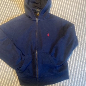 Marinblå hoodie från Polo Ralph Lauren - Säljer en marinblå hoodie från Polo Ralph Lauren med dragkedja och röd broderad logga på bröstet. Jackan har huva och två fickor framtill. Perfekt för en avslappnad stil.