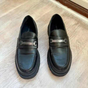 Svarta loafers från Pull&Bear - Stilrena svarta loafers från Pull&Bear med metallspänne över vristen. Skorna har rund tå och en rejäl platt sula som ger en modern look.