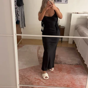 Svart långklänning i satin - Säljer en elegant svart långklänning i satin med smala axelband och figurnära passform. Klänningen har en stilren design och en lätt glansig yta som ger en lyxig känsla. Perfekt för dig som vill ha en klassisk och tidlös look.