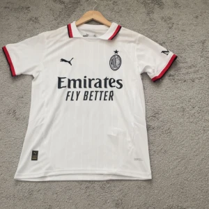 AC Milan vit bortatröja Puma - Säljer en vit AC Milan bortatröja från Puma med svarta och röda detaljer på krage och ärmslut. Tröjan har klubbmärke och sponsortryck på bröstet samt diskreta vertikala ränder. Perfekt för fotbollsfans!