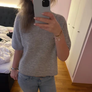 Grå stickad kortärmad tröja - Mysig grå stickad tröja med korta ärmar och rund hals. Tröjan har en enkel och stilren design med ribbade kanter vid ärmslut och nederkant. Perfekt för lager-på-lager eller sommarens svalare dagar.