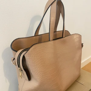 Beige axelväska med krokodilpräglat mönster - Säljer en beige axelväska med krokodilpräglat mönster och svarta kanter. Väskan har dubbla handtag och en stor dragkedjeförsedd innerficka. Perfekt för att bära med sig allt man behöver till vardags.