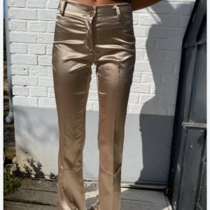 Guldiga satinbyxor   - Snygga beiga/guldiga byxor i satin med glansig finish och utsvängda ben. Byxorna har låg/medel midja, dragkedja och knapp framtill.