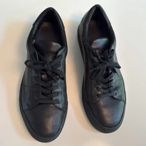 Svarta sneakers från DNDP - Säljer ett par helsvarta sneakers från DNDP med perforerat mönster och snörning. Skorna har rund tå och stilren look. Perfekta för dig som gillar minimalistisk stil.