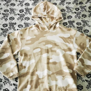 Beige camouflagemönstrad hoodie från Lindex - Säljer en beige och vit camouflagemönstrad hoodie från Lindex. Tröjan har huva och långa ärmar, perfekt för dig som gillar en avslappnad stil. Mjuk insida och elastiska muddar vid ärmslut och nederkant.