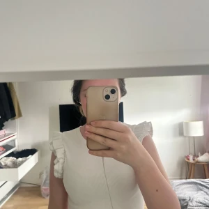 Vit ribbad topp med volangärmar från Zara - Säljer en vit ribbad topp från Zara med korta volangärmar. Toppen har rund halsringning och är tillverkad i ett stretchigt material som sitter snyggt på kroppen. Perfekt för en fräsch och stilren look.