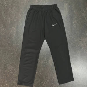 Svarta träningsbyxor från Nike - Svarta träningsbyxor från Nike med elastisk midja och två sidofickor. Byxorna har en rak passform och är tillverkade i ett lätt syntetmaterial. Perfekta för sport eller avslappnade dagar. Nike-logga i vitt på låret.