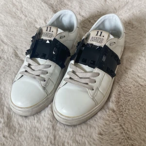 Valentino Rockstud Untitled - Säljer ett par vita Valentino Rockstud Untitled sneakers med svarta nitar och bred svart rem över vristen. Skorna har snörning, rund tå och är tillverkade i skinn. Klassisk och edgy design, Valentino märket på sulan har gått bort när man använd dem, men syns fortfarande pyttelite. Man får med 4 nitar men ingen box eller annat då jag köpt de på Plick och fick inte med, skriv för frågor 🥰 är inte helt säker på storlek