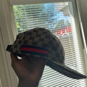 Beige keps från Gucci med monogram - Säljer en beige keps från Gucci med klassiskt GG-monogram och röd-blå rand på sidan. Kepsen har böjd skärm och justerbar rem baktill. Perfekt accessoar för en trendig look.