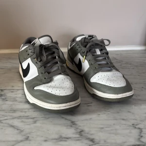 Nike Dunk Low NBA sneakers i grått och vitt - Knappt använda! Nike Dunk Low NBA sneakers i grått och vitt med svart swoosh. Skorna har snörning, rund tå och platt sula. NBA-detalj på sidan och Nike-logga bak. Perfekta för dig som gillar streetwear och sportig stil.