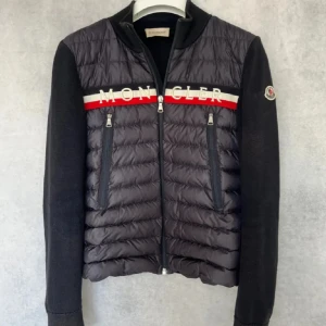 Svart hybridjacka från Moncler - Säljer en svart Moncler-jacka med quiltad front och huva, stickade ärmar och sidor. Jackan har dragkedja framtill, två fickor med dragkedja och Moncler-logga på ärmen. Snygg detalj med röd och vit rand över bröstet samt broderad Moncler-text.