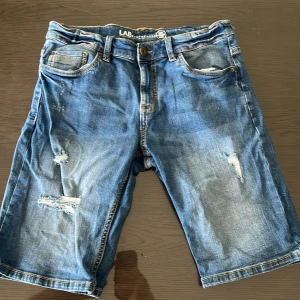 Jeansshorts från LAB Industries - Snygga blå jeansshorts från LAB Industries med slitna detaljer och klassisk femficksdesign. Shorts i denim med knapp och dragkedja framtill, samt patch bak i midjan. Perfekta för en avslappnad stil.