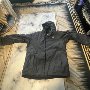 Svart vindjacka från The North Face - Säljer en svart vindjacka från The North Face med huva och dragkedja. Jackan har två sidofickor och klassisk logga på bröstet. Perfekt för blåsiga dagar och enkel att matcha med olika outfits.