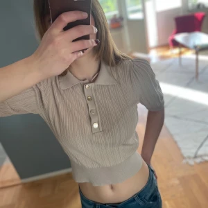 Beige ribbad pikétröja med knappar - Säljer en beige ribbad pikétröja med krage och tre knappar framtill. Tröjan har kort ärm och är croppad i modellen. Finns en tråddragning på ena ärmen. Lindex Holly & White. 