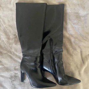 Svarta höga stövlar med klack - Säljer ett par svarta höga stövlar med spetsig tå och klack. Skorna har dragkedja på insidan och är tillverkade i ett glansigt material som liknar skinn. Perfekta för att lyfta vilken outfit som helst. Jag vet inte vad storleken är. Jag själv har 38 i klackskor och den är lite för stor så antar 39.