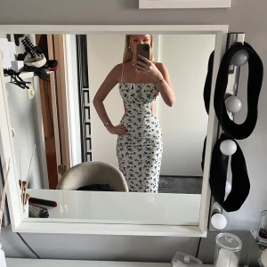 Vit långklänning med blå blommor - Säljer en vit långklänning med blått blommönster och smala axelband. Klänningen har tight passform, öppen rygg, använd 2 gånger max, går ner till fötterna på mig som är 168