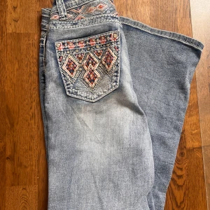 Blå bootcut jeans med broderad bakficka - Snygga blå bootcut jeans med broderad bakficka i rosa, orange och vitt samt nitar. Jeansen har lågmidjad passform och klassisk femficksdesign. Aldrig använd då jag redan har ett par!💗