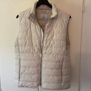 Beige dunväst från Bläck - Stilren beige dunväst från Bläck med ståkrage och dragkedja framtill. Västen har ett quiltat mönster och är i toppskick. Säljer eftersom att den blivit för liten för mig. Strl M. Passar perfekt nu under våren. Om du har några funderingar tveka inte att skriva!!