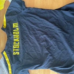 AIK t shirt  - Svart t-shirt med gula detaljer och texten 'STOCKHOLM' på ryggen. AIK-logga på bröstet. Klassisk passform och korta ärmar, perfekt för dig som vill visa ditt AIK-stöd.