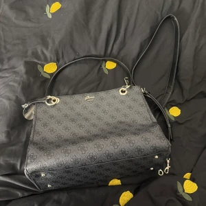 Svart axelväska från Guess med mönster - Snygg svart axelväska från Guess med diskret logomönster och guldfärgade metalldetaljer. Väskan har både handtag och axelrem samt dragkedja upptill. Insidan är ljus med flera fack för enkel organisering.