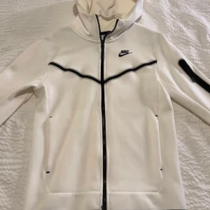 Vit tracksuit från Nike  - Säljer en vit tracksuit från Nike med svarta dragsnören och logga på vänster lår. Byxorna har resår i midjan, fickor på sidorna och en svart dragkedjedetalj på ena benet. Perfekta för en sportig och avslappnad stil.