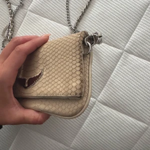 Beige axelväska med kedjerem och ormskinnsmönster - Säljer en beige axelväska från zadig med snyggt ormskinnsmönster och silverfärgad kedjerem. Väskan har dubbla dragkedjor och en ikonisk silverfärgad fågeldetalj på framsidan. Perfekt för dig som vill ha en stilren accessoar med det lilla extra. Den har lite defekter på sidan och kedjan, visas på bilderna.