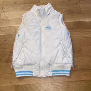 Vit dunväst från Happy Girl - Vit dunväst med ljusblå detaljer och broderad logga från Happy Girl. Västen har dragkedja framtill, ribbade muddar med blå ränder och två fickor med dragkedja. Perfekt för lager-på-lager under kyliga dagar.