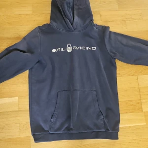 Mörkblå hoodie från Sail Racing 170 - Säljer en mörkblå hoodie från Sail Racing med vit logga på bröstet. Tröjan har huva och en stor magficka framtill. Perfekt för dig som gillar en sportig och stilren look.