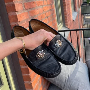 Svarta loafers från Ralph Lauren - Säljer ett par klassiska svarta loafers från Ralph Lauren med RL-logga i silver framtill och röd RL-detalj bak. Skorna har rund tå och är tillverkade i skinn med en platt sula och diskret söm längs kanterna. Använt 5 gånger. Nypris 1700kr.