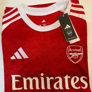 Arsenal hemmatröja 2023/24 - Alla storlekar finns Röd och vit Arsenal hemmatröja från Adidas med klubbmärke och sponsortryck på bröstet. Tröjan har vita detaljer på axlar och ärmar samt ett subtilt mönster med bokstaven A över hela framsidan. Rund halsringning och klassisk passform.