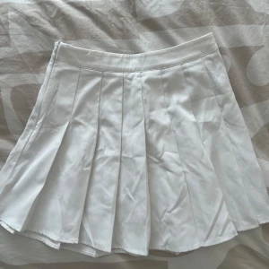 Vit plisserad kortkjol - Säljer en vit plisserad kortkjol med klassisk tennisstil. Kjolen har bred linning och tydliga veck som ger en snygg och sportig look. Perfekt att matcha med en enkel topp eller hoodie.
