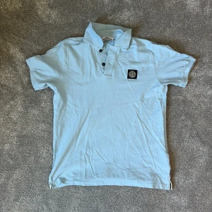 Ljusblå pikétröja från Stone Island Junior - Säljer en ljusblå pikétröja från Stone Island Junior med klassisk krage och knappslå framtill. Tröjan har korta ärmar och en broderad logotyp på bröstet. Perfekt för en avslappnad och stilren look.