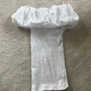 Vit offshoulder klänning med puffärm - Trendig vit klänning med stor puffig offshoulder-ärm och rak passform. Perfekt för dig som vill sticka ut med en unik siluett. Klänningen är axelbandslös och har en minimalistisk design som ger en modern känsla.