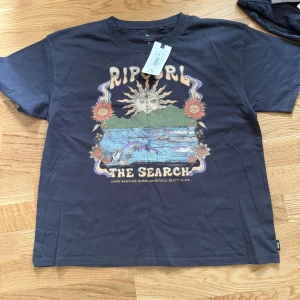Svart t-shirt från Rip Curl med tryck - Ny, Cool svart t-shirt från Rip Curl med färgglatt tryck framtill som visar sol, blommor och vågor. T-shirten har en klassisk passform och rund halsringning. Perfekt för dig som gillar surf och avslappnad stil.