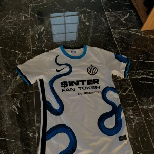 Inter Milan vit bortatröja 2021/22 - Inter Milans bortatröja från säsongen 2021/22. Vit med blå och svart orm-mönster, svart Nike-logga och klubbmärke på bröstet. Blå detaljer vid krage och ärmslut. Sponsortryck på framsidan.