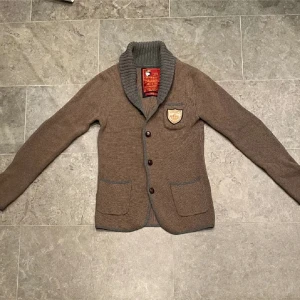Morris cardigan - Säljer en brun stickad kofta från Morris. Med ett snyggt emblem på bröstet. Perfekt för kyliga dagar!