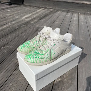 Maison Margiela sneakers med grön färgstänk - Unika sneakers från Maison Margiela i vitt skinn med coola gröna färgstänk. De har en klassisk snörning och en bekväm passform. Perfekta för dig som vill sticka ut med en konstnärlig touch på dina skor. Använda men i bra skick, redo för nya äventyr!
