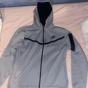 Grå hoodie jacka från Nike - Snygg grå hoodie jacka från Nike med svart logga på bröstet och svarta detaljer över bröstet och på ena ärmen. Jackan har dragkedja hela vägen och huva. Perfekt för en sportig look.