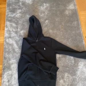 Svart hoodie från Polo Ralph Lauren - Säljer en svart hoodie från Polo Ralph Lauren med dragkedja och huva. Väldigt bra skick, bara använd några gånger. Passar ungefär mellan 165cm - 175cm