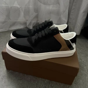 Burberry skor - Snygga svarta sneakers från Burberry med klassiskt rutigt mönster i beige, svart och vitt på hälen. Skorna är nästan helt oanvända på grund av att de passar inte mig. Pris kan diskuteras! Vid fler frågor eller funderingar så är det bara att höra av sig!😄