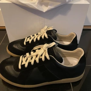 Svarta sneakers med vita snören från Maison Margiela - Säljer ett par svarta sneakers från Maison Margiela med vita snören och beige sula. Skorna har en klassisk design med rund tå och paneler i skinn och mocka. Perfekta för dig som gillar stilrena och tidlösa sneakers. Kvitto och Box finns 