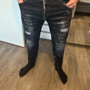 Svarta jeansbyxor från Dsquared2 - Säljer ett par svarta jeansbyxor från Dsquared2 med slitningar och distressed detaljer framtill. Byxorna har smal passform och klassisk femficksdesign. Märkeslapp bak med texten 'IBRA BLACK ON BLACK DSQUARED2'. 