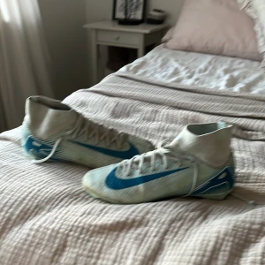 Vita och blåa fotbollsskor från Nike - Säljer ett par vita och blåa Nike fotbollsskor med snörning och rund tå. Skorna har dobbar för spel på gräs och en sportig design med Nike-logga på sidan. Perfekta för dig som vill ta ditt spel till nästa nivå.