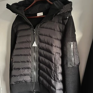 Svart hybridjacka från Moncler - Säljer en svart Moncler-jacka med vadderad front och stickade ärmar. Jackan har huva, dragkedja framtill och en ficka med dragkedja på ärmen. Klassisk Moncler-logga på armen. Perfekt för kyliga dagar.