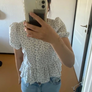 Vit blommig blus från KappAhl - Supersöt vit blus med små blå blommor från KappAhl. Blusen har puffiga korta ärmar, volang nertill och knappar framtill. Perfekt för vår och sommar, lätt och luftig i bomullsmaterial.