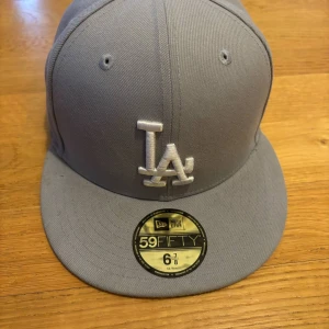 Grå LA-keps från New Era 59FIFTY - Snygg grå keps från New Era 59FIFTY med LA-logga framtill i vitt. Klassisk modell med rak skärm och broderade ventilationshål. Tillverkad i polyester och passar perfekt för dig som gillar sportig stil. Bara en liten fläck på skärmen som nästan inte ens syns.
