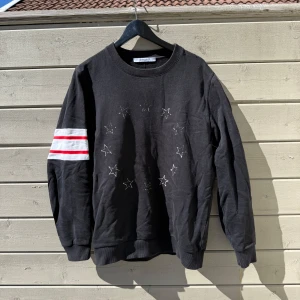 Svart sweatshirt från Givenchy med stjärnmönster - Säljer en svart sweatshirt från Givenchy med broderade vita stjärnor på bröstet och två röda ränder på ena ärmen. Tröjan har rund halsringning och ribbade muddar vid ärmslut och nederkant. Köpt ifrån Viaggio Club, så garanterat äkta!