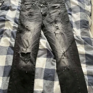 Svarta slitna jeans från Dsquared2 - Säljer ett par svarta jeans från Dsquared2 med slitningar och hål på benen. Jeansen har klassisk femficksdesign, dragkedja och knapp i midjan. Snyggt tvättade detaljer och Dsquared2-logga vid gylfen samt bak på linningen.